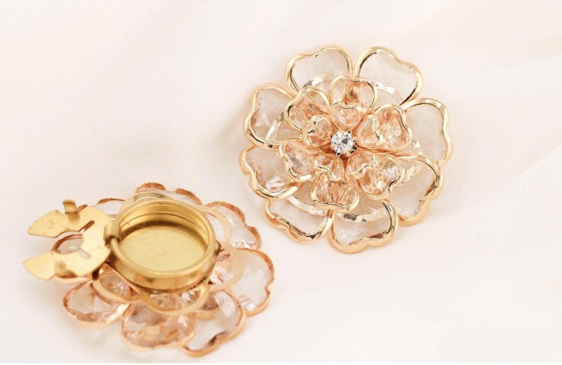 May include: Two flower-shaped decorative objects. The flowers have a gold-coloured metal frame with clear, faceted petals. One flower has a small, round, gold-coloured base. The other flower has a small, clear, round gem in the centre.