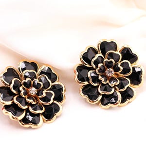 May include: A pair of black flower-shaped earrings with gold-coloured trim. Each earring features a central crystal and multiple black, faceted petals. The earrings are set against a white background.