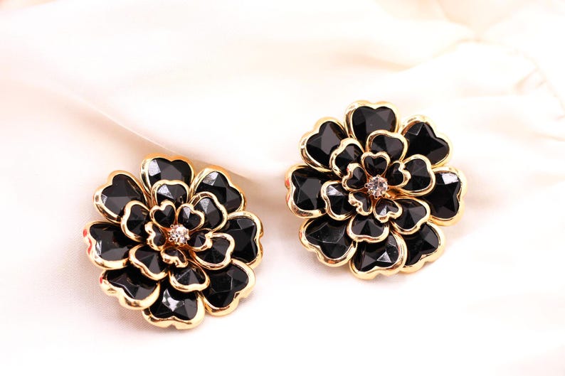 May include: A pair of black flower-shaped earrings with gold-coloured trim. Each earring features a central crystal and multiple black, faceted petals. The earrings are set against a white background.