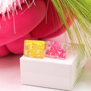 May include: Two square resin rings, one with yellow beads and the other with pink beads, sit on a white box. A pink plush object and green decorative grass are in the background. The rings are a unique fashion accessory.