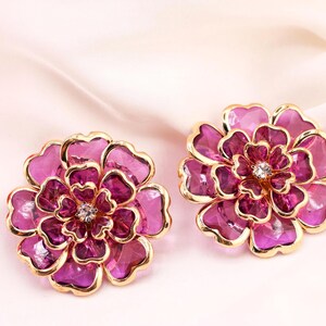 May include: A pair of flower-shaped earrings with gold-coloured edging and a central crystal. The petals are a deep magenta with a lighter pink shade. The earrings are displayed on a soft, light pink background.