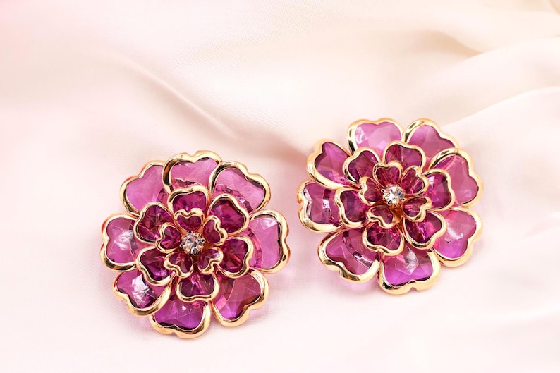 May include: A pair of flower-shaped earrings with gold-coloured edging and a central crystal. The petals are a deep magenta with a lighter pink shade. The earrings are displayed on a soft, light pink background.