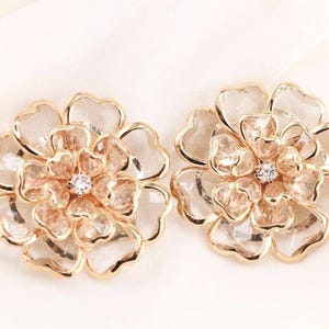 May include: A pair of gold-coloured flower-shaped earrings. Each earring has a central clear stone, surrounded by petals with a clear, faceted appearance. The earrings are set against a white background.