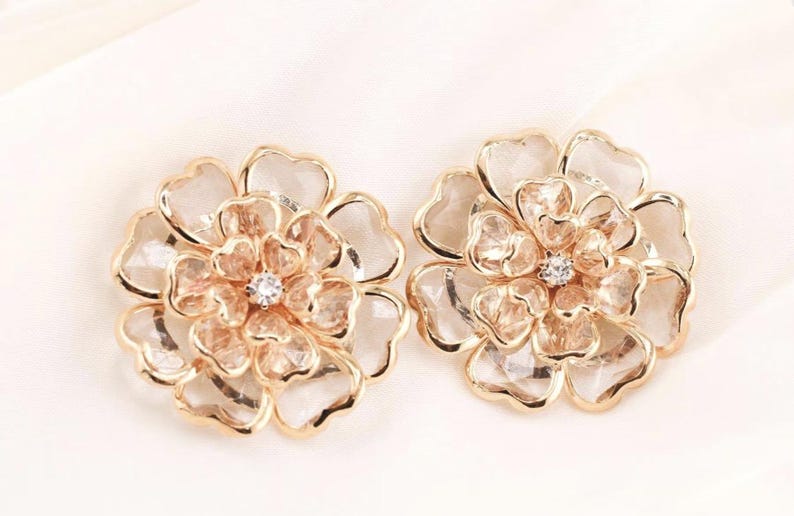 May include: A pair of gold-coloured flower-shaped earrings. Each earring has a central clear stone, surrounded by petals with a clear, faceted appearance. The earrings are set against a white background.