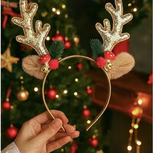 May include: A gold-colored headband with reindeer antlers covered in gold glitter. The antlers are adorned with small red pom-poms, faux fur, and tiny bells. The headband is a festive accessory for the holiday season.