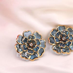 May include: A pair of flower-shaped earrings with gold-coloured edging. The earrings are a blue-green colour and feature a central crystal. They are displayed on a white, silky background.