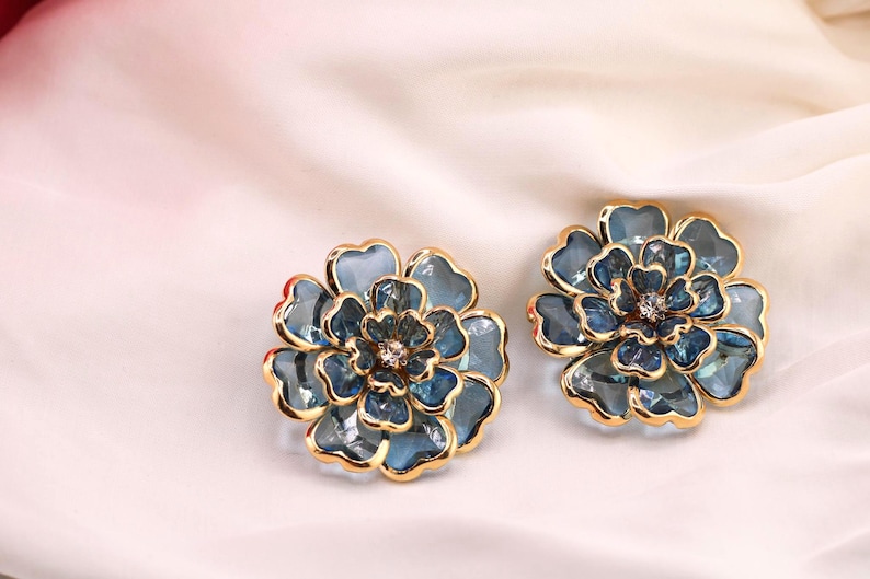 May include: A pair of flower-shaped earrings with gold-coloured edging. The earrings are a blue-green colour and feature a central crystal. They are displayed on a white, silky background.