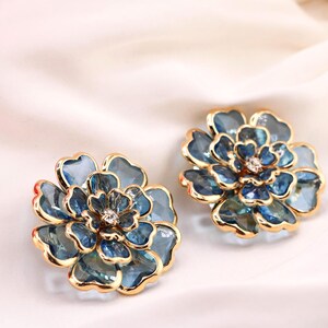 May include: A pair of flower-shaped earrings. Each earring has a blue and gold design, with a central crystal. The petals display a gradient of blue shades. The earrings are set against a white, silky background.