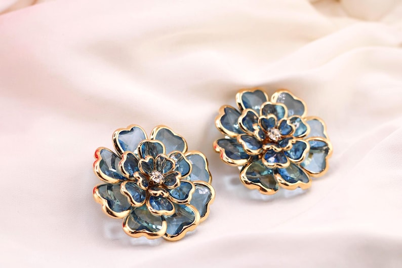 May include: A pair of flower-shaped earrings. Each earring has a blue and gold design, with a central crystal. The petals display a gradient of blue shades. The earrings are set against a white, silky background.