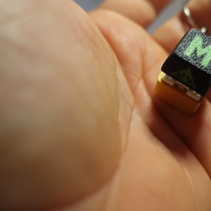 May include: A black and yellow key chain with a green letter "M" on the front. The keychain is held in a hand.