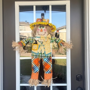 May include: A fabric scarecrow door hanging featuring a hat with yellow pom-poms, a green plaid shirt, a yellow patterned vest, and orange plaid pants. The figure has raffia hands and feet, a painted face, and small black bird accents.