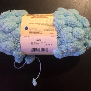 May include: A ball of Capri yarn in a light blue color. The yarn has a textured, pom-pom-like appearance. The label reads "CAPRI" and includes the color code 9806 and dye lot number 121700. The brand is Coats & Clark.