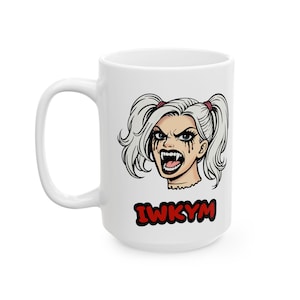 以下が含まれることがあります： 白いセラミックマグ。白いおさげ髪、黒いアイメイク、牙を持つ女性の漫画イラストが描かれています。マグには赤い文字で「IWKYM」と書かれています。