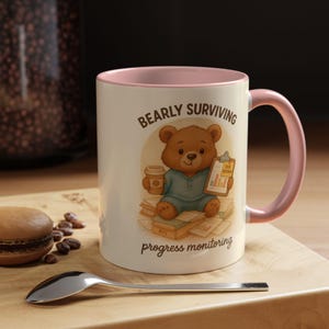 May include: A white ceramic mug with a pink interior and handle. The mug features a cartoon bear illustration with the text "BEARLY SURVIVING progress monitoring". The bear is holding a coffee cup and a clipboard with charts.