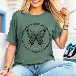 May include: Sage green t-shirt featuring a detailed black butterfly design within a circular text frame. The text includes phrases like "Babe-Glow Mode" and "Pretty Power-Boss". The shirt is a casual, everyday wear item.