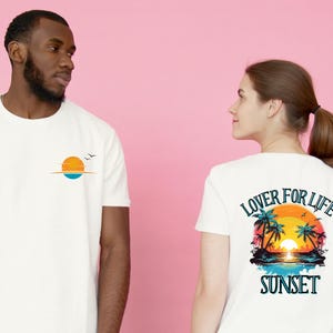 Peut inclure: Un t-shirt blanc à manches courtes avec un petit motif de coucher de soleil et d'oiseaux sur la poitrine. Le dos du t-shirt présente une grande scène tropicale colorée avec des palmiers, de l'eau et le texte LOVER FOR LIFE SUNSET.