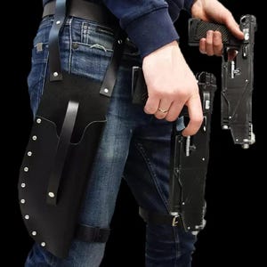 May include: Black leather thigh holster with riveted edges and a belt loop. The holster is worn with a black belt and a leg strap. A person is holding two black futuristic-looking firearms.