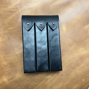 May include: A black leather pouch with three vertical compartments. The top flap is secured with three brass-coloured rivets. The pouch is rectangular and appears to be handmade, with a focus on functionality and durability.