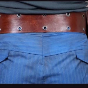 May include: A wide brown leather utility belt featuring two vertical side pouches and two horizontal rows of silver-toned metal grommets. The belt is worn over blue trousers and a grey shirt, showcasing a structured design with visible stitching.