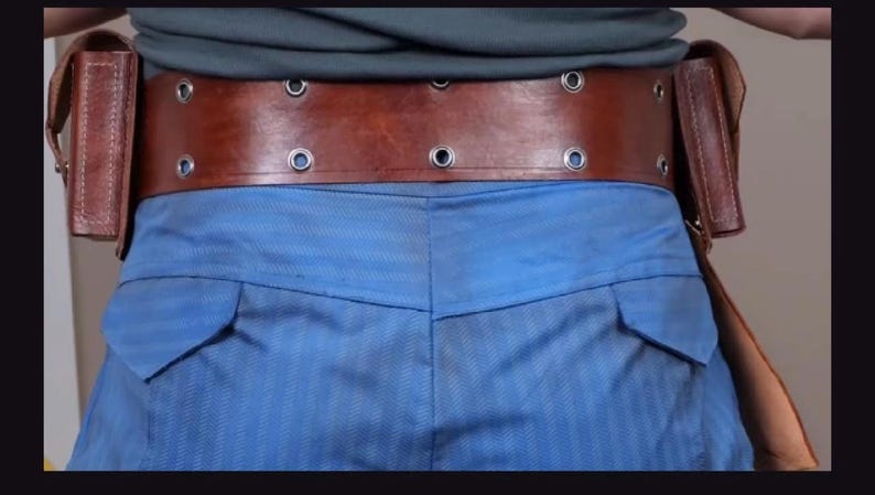 May include: A wide brown leather utility belt featuring two vertical side pouches and two horizontal rows of silver-toned metal grommets. The belt is worn over blue trousers and a grey shirt, showcasing a structured design with visible stitching.