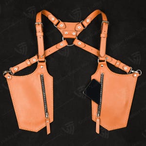 May include: A tan leather chest holster with two zippered pockets and adjustable straps. The holster has a harness design with metal hardware and is designed to carry a phone. The product is made of leather.