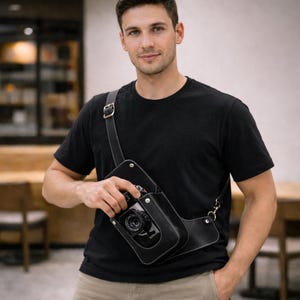 Puede incluir: Bolso de cámara de cuero negro con costuras blancas, usado cruzado. El bolso contiene una cámara negra. La correa es ajustable con una hebilla de metal. El bolso tiene un bolsillo lateral. La persona lleva una camiseta negra.
