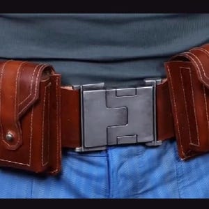 May include: A brown leather utility belt featuring a large, rectangular, brushed metal buckle at the centre. The belt includes four attached vertical pouches with flap closures, each secured with a small metal stud, and visible white stitching.