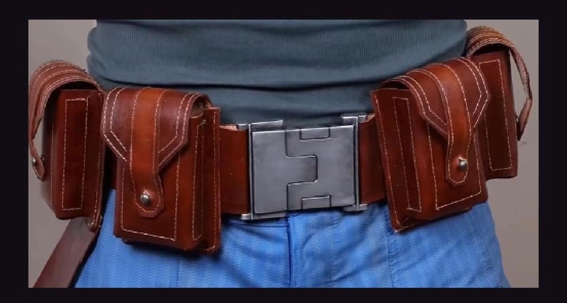 May include: A brown leather utility belt featuring a large, rectangular, brushed metal buckle at the centre. The belt includes four attached vertical pouches with flap closures, each secured with a small metal stud, and visible white stitching.