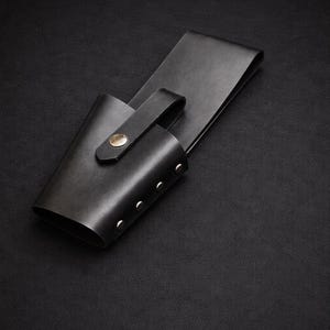 May include: A black leather sheath with a belt loop and a strap secured by a gold-colored rivet. The sheath has a cylindrical pocket with three silver rivets along the side. The background is a dark, textured surface.