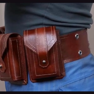 May include: A brown leather utility belt featuring two rectangular pouches with flap closures secured by metal snaps. The belt is constructed with visible stitching along the edges and includes two metal grommets on the side strap section.