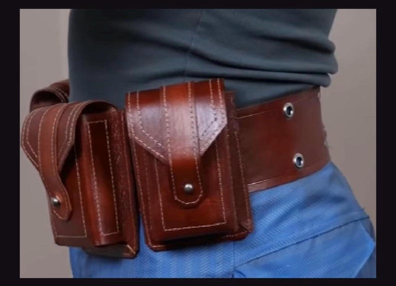 May include: A brown leather utility belt featuring two rectangular pouches with flap closures secured by metal snaps. The belt is constructed with visible stitching along the edges and includes two metal grommets on the side strap section.