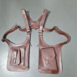 May include: A brown leather shoulder holster with two pouches, zippers, and adjustable straps. The holster has a harness design with a connecting piece at the back and adjustable straps. The leather is a rich, medium brown color.