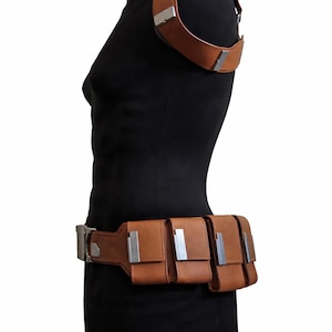 May include: Brown leather shoulder strap and belt set with silver-colored metal accents. The belt features three pouches. The set is displayed on a black mannequin.