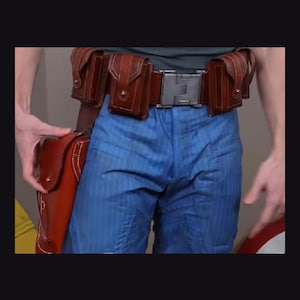May include: A brown leather utility belt featuring multiple small pouches and a large holster attached to the side, worn over blue trousers with a subtle vertical stripe pattern. The belt is secured with a metallic rectangular buckle at the centre.
