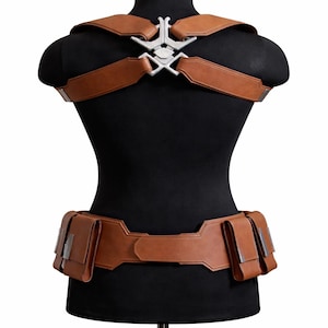 May include: Brown leather harness and belt set with silver-colored metal accents. The harness features shoulder straps connected by a decorative piece. The belt has multiple pouches. The set is displayed on a black mannequin.