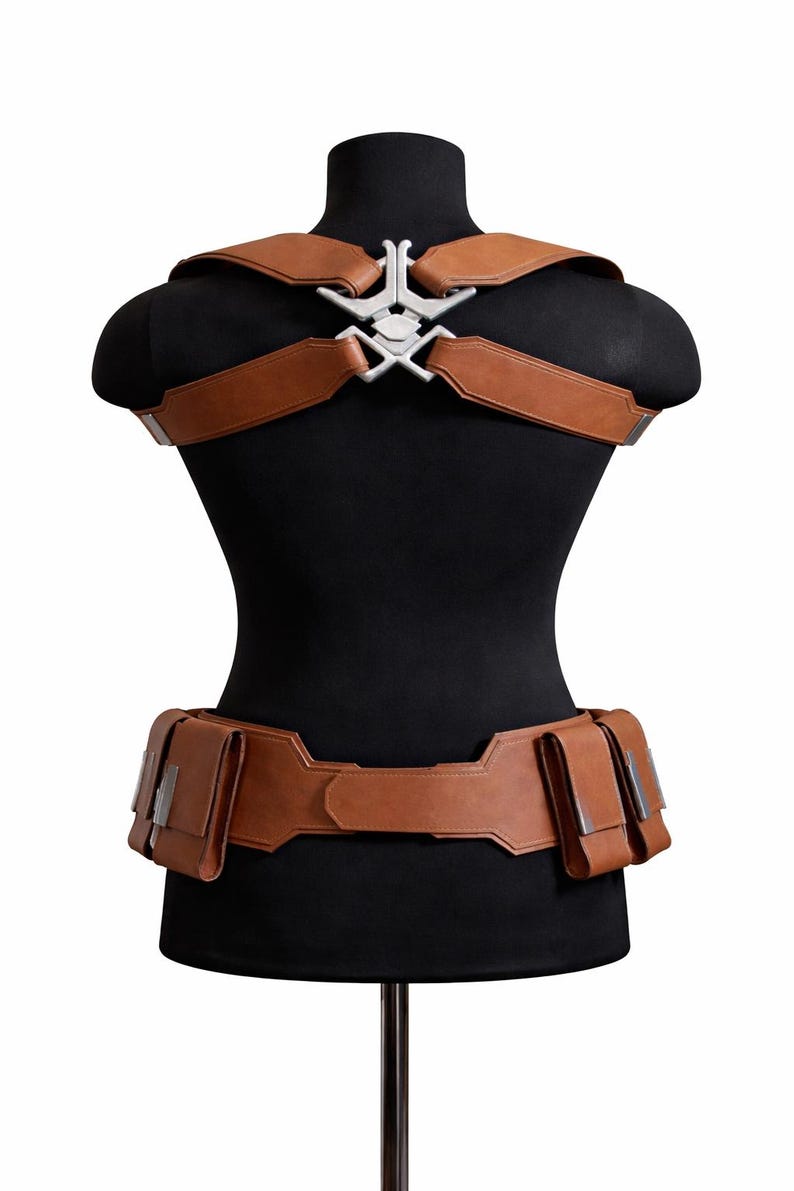 May include: Brown leather harness and belt set with silver-colored metal accents. The harness features shoulder straps connected by a decorative piece. The belt has multiple pouches. The set is displayed on a black mannequin.