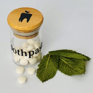 Puede incluir: Un frasco de vidrio transparente lleno de tabletas de pasta de dientes blancas, con la palabra "toothpaste" impresa en el lateral. El frasco tiene una tapa de madera con un gr&aacute;fico de diente negro. Hojas de menta verde fresca est&aacute;n al lado del frasco.