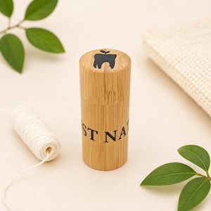 Puede incluir: Un recipiente de bambú con un logotipo negro de diente y manzana en la parte superior, y el texto "ST NA" en el lateral. También se ven un carrete de hilo dental blanco y hojas verdes.