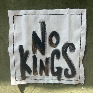 May include: A white fabric patch with black stitching around the edges. The words "No Kings" are painted in black and gold on the patch.