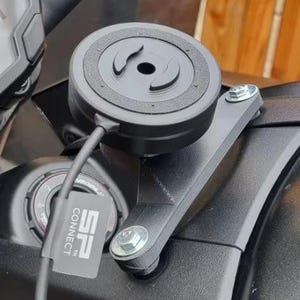Op de afbeelding: Zwart motorfietsdashboard met een SP Connect telefoonhouder bevestigd. De houder is cirkelvormig en verbonden met een beugel. De motorfiets heeft een digitaal display en zwarte stuur. De achtergrond is een houten hek.