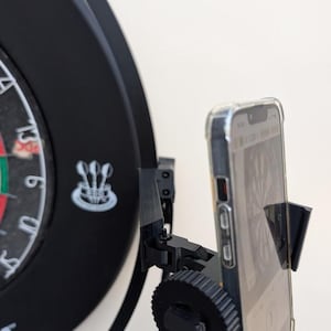Op de afbeelding: Een zwarte plastic smartphonehouder bevestigd aan de zijkant van een rond dartbord. De houder is voorzien van een verstelbare knop en een houder voor een smartphone met een doorzichtig beschermhoesje, gericht op de voorzijde van het dartbord.