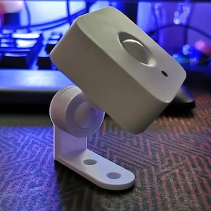 Op de afbeelding: Een wit, rechthoekig apparaat met een afgeronde bovenkant en een cirkelvormige inkeping. Het is gemonteerd op een witte beugel met een cirkelvormig gewricht en een basis met twee gaten. Het apparaat is schuin geplaatst, wat suggereert dat het een camera of sensor is.