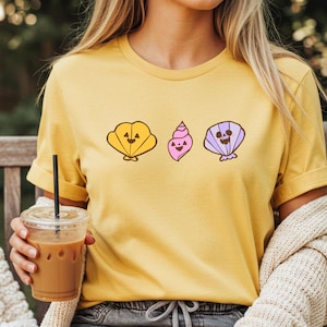 May include: Yellow t-shirt featuring three cartoon seashell designs in yellow, pink, and purple. The shirt is made of a soft material and is suitable for casual wear. The design is simple and eye-catching.