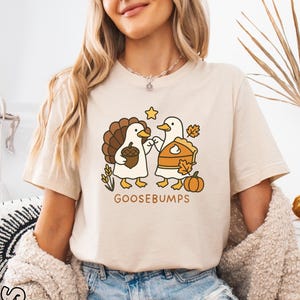 May include: A soft cream-colored t-shirt featuring a cartoon illustration of two ducks dressed as a turkey and a pumpkin pie. The text "GOOSEBUMPS" is printed below the ducks. The shirt is worn by a person wearing denim shorts and a cream-colored cardigan.