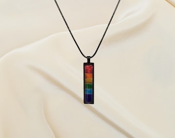 May include: A black necklace with a rectangular pendant featuring a rainbow design. The pendant has a black border and displays the colors of the rainbow in vertical stripes. The necklace hangs against a white, silky background.