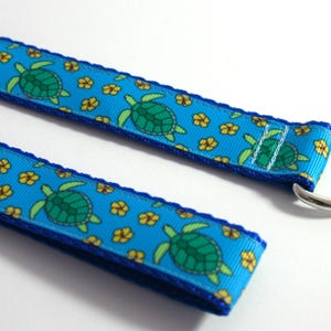 May include: A turquoise ribbon featuring a pattern of green turtles and yellow flowers. The ribbon has a dark blue border and a silver metal ring attached to one end. Likely for a pet collar or lead, the ribbon is approximately 2.5 cm wide.
