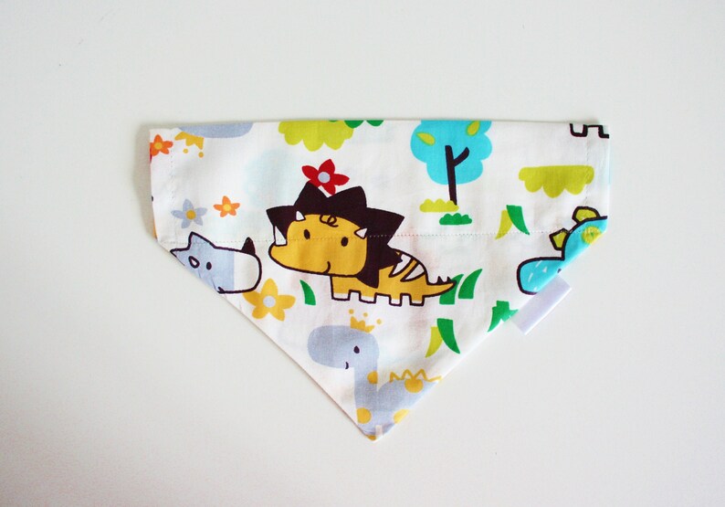 May include: A white cotton dog bandana with a colorful dinosaur print. The bandana features a yellow dinosaur with a red flower, a blue dinosaur, and a gray rhino. The bandana is trimmed with a white border.