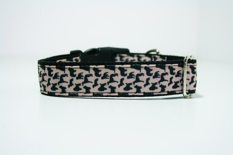 May include: A black and pink dog collar with a repeating pattern of black silhouettes of dogs. The collar has a black plastic buckle and a silver metal D-ring.