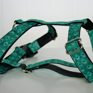 Puede incluir: Un arnés para perro con estampado de plumas de pavo real verde y azul, con correas negras y hebillas plateadas.