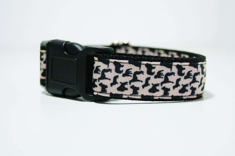 May include: A black dog silhouette pattern on a pink and black dog collar with a black plastic buckle.